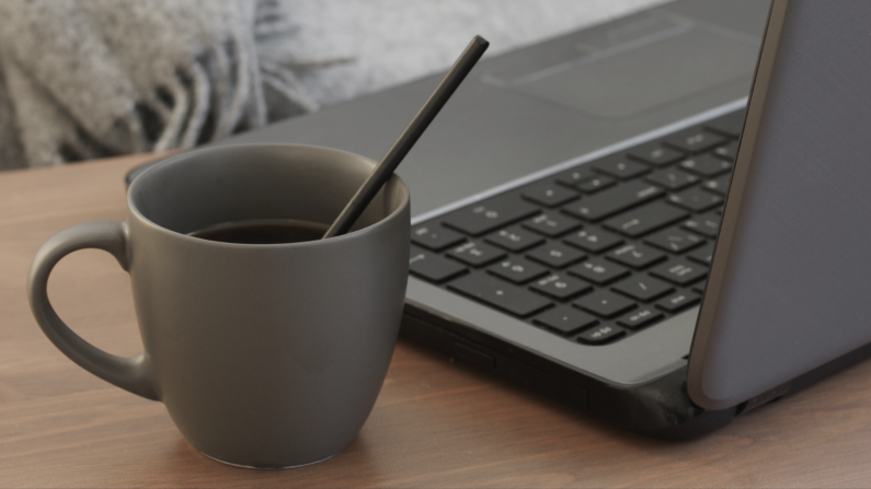 laptop, obok szary kubek z kawą, w tle kanapa i koc, szare